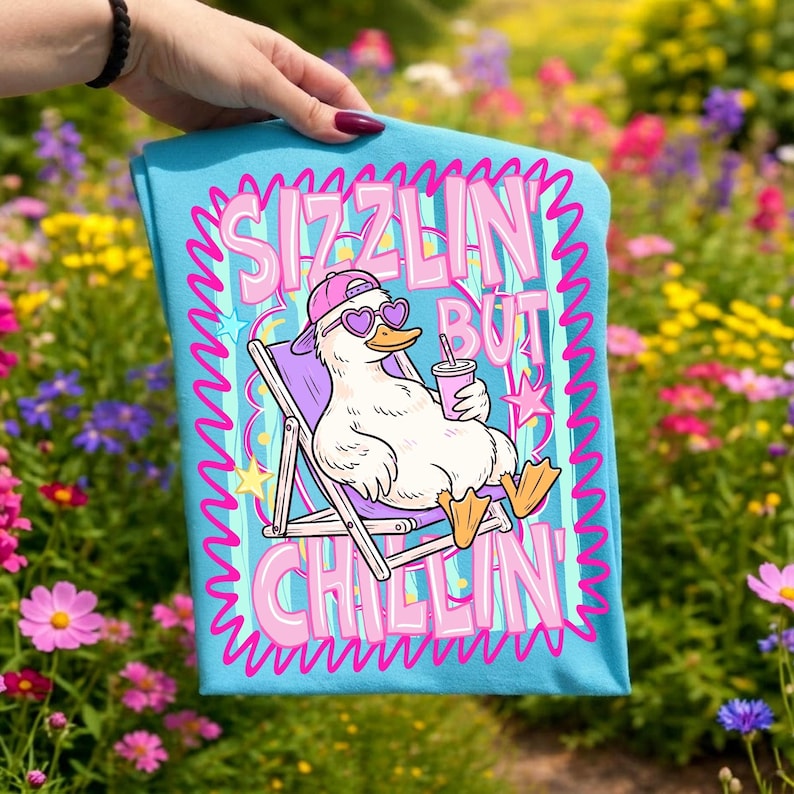 Op de afbeelding: Een lichtblauw t-shirt met een cartoon eend met een hartvormige zonnebril en een roze pet, ontspannen in een strandstoel met een drankje. Het shirt heeft de tekst "Sizzlin' But Chillin'" in roze en wit.