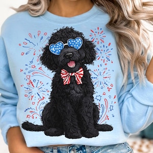 Pu&ograve; includere: Felpa azzurra con un barboncino nero che indossa occhiali da sole a forma di cuore con un design a stelle e strisce e un papillon rosso, bianco e blu. Il design include fuochi d'artificio rossi, bianchi e blu.