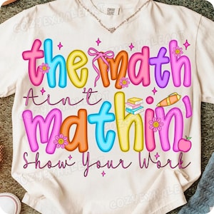 May include: Cream-colored t-shirt with a colorful graphic that reads "the math ain't mathin' Show Your Work." The text is in various colors, including pink, blue, yellow, and orange, with flower and star accents. A fun, educational tee.