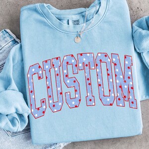 Op de afbeelding: Een lichtblauw sweatshirt met het woord "CUSTOM" in grote, vette letters over de voorkant. De letters zijn rood omlijnd en gevuld met een patroon van witte en rode sterren. Een zilveren ketting met een kleine hanger is zichtbaar.