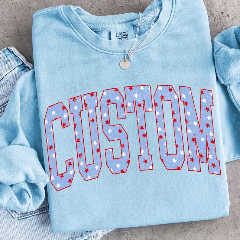 Op de afbeelding: Een lichtblauw sweatshirt met het woord "CUSTOM" in grote, vette letters over de voorkant. De letters zijn rood omlijnd en gevuld met een patroon van witte en rode sterren. Een zilveren ketting met een kleine hanger is zichtbaar.