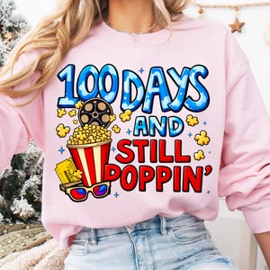 Puede incluir: Sudadera rosa claro con el texto "100 DAYS AND STILL POPPIN'" en azul y rojo. El dise&ntilde;o incluye una bobina de pel&iacute;cula, palomitas de ma&iacute;z, una entrada de cine y gafas 3D. Un estilo casual y c&oacute;modo.