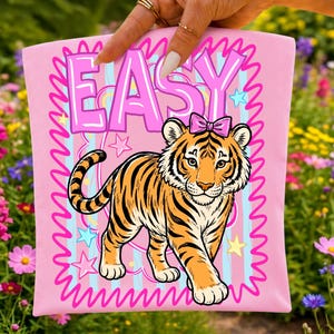 May include: A pink t-shirt featuring the word "EASY" in a bubble font. Below the text is a cartoon tiger with orange and black stripes and a pink bow. The design is framed by a pink, wavy border with stars.