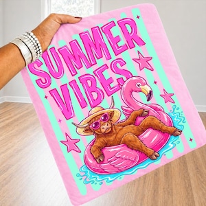 Może przedstawiać: R&oacute;żowy koc z napisem "SUMMER VIBES" w jasnor&oacute;żowym kolorze. Koc przedstawia kresk&oacute;wkową ilustrację brązowej krowy w okularach przeciwsłonecznych i słomkowym kapeluszu, relaksującej się na r&oacute;żowym pływaku w kształcie flaminga. R&oacute;żowe gwiazdki i turkusowe paski dopełniają projekt.