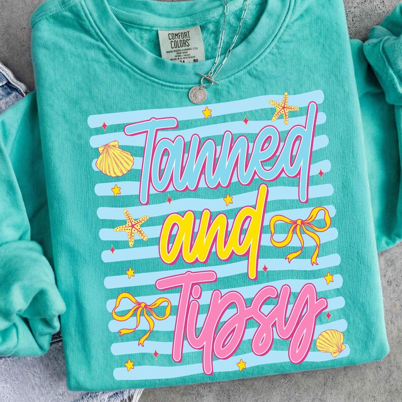 Peut inclure: Sweat-shirt Comfort Colors turquoise avec l'inscription &laquo; Tanned and Tipsy &raquo; en bleu, jaune et rose. Le motif comprend des coquillages, des &eacute;toiles de mer et des n&oelig;uds. Un collier argent&eacute; avec un pendentif est visible.