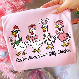 May include: Pink t-shirt featuring a cartoon design of four chickens dressed for Easter. One chicken wears sunglasses and a bandana, another a bow, a third has bunny ears and glasses, and the last a sweater and hat. Text reads "Easter Vibes, Same Silly Chicken."
