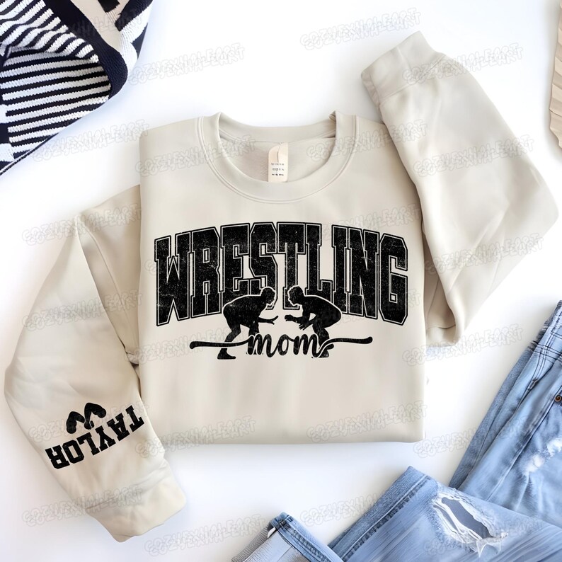 May include: A cream-colored sweatshirt with the words "WRESTLING mom" in black block letters and a graphic of two wrestlers. The left sleeve has the name "TAYLOR" printed in black. The sweatshirt is paired with blue jeans.
