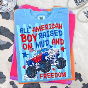 K&ouml;nnte beinhalten: Ein hellblaues Comfort Colors T-Shirt mit dem Text "ALL AMERICAN BOY RAISED ON MUD AND FREEDOM" und einer Grafik eines rot, wei&szlig; und blauen Monster Trucks. Das T-Shirt ist auf anderen bunten Shirts gefaltet.