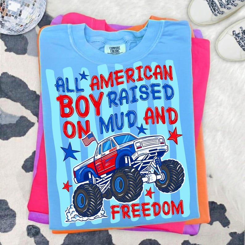 K&ouml;nnte beinhalten: Ein hellblaues Comfort Colors T-Shirt mit dem Text "ALL AMERICAN BOY RAISED ON MUD AND FREEDOM" und einer Grafik eines rot, wei&szlig; und blauen Monster Trucks. Das T-Shirt ist auf anderen bunten Shirts gefaltet.