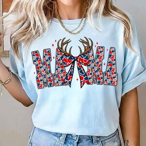Pu&ograve; includere: T-shirt azzurra con la scritta "MAMA" in un motivo mimetico rosso, bianco e blu. Un fiocco con lo stesso motivo e corna di cervo sono sopra la parola. La maglietta &egrave; in stile casual.