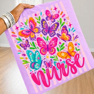 May include: A light purple t-shirt featuring a vertical striped pattern and a colorful graphic print of multiple butterflies in shades of pink, orange, yellow, and teal, accented with green leaves and the word Nurse in a bright pink script font.