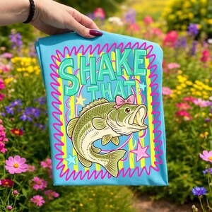 Op de afbeelding: Lichtblauw t-shirt met een cartoon bas met een roze strik en de tekst "SHAKE THAT". Het ontwerp is omlijst met een golvende roze rand en gele strepen. Het t-shirt wordt gepresenteerd voor een kleurrijke bloementuin.
