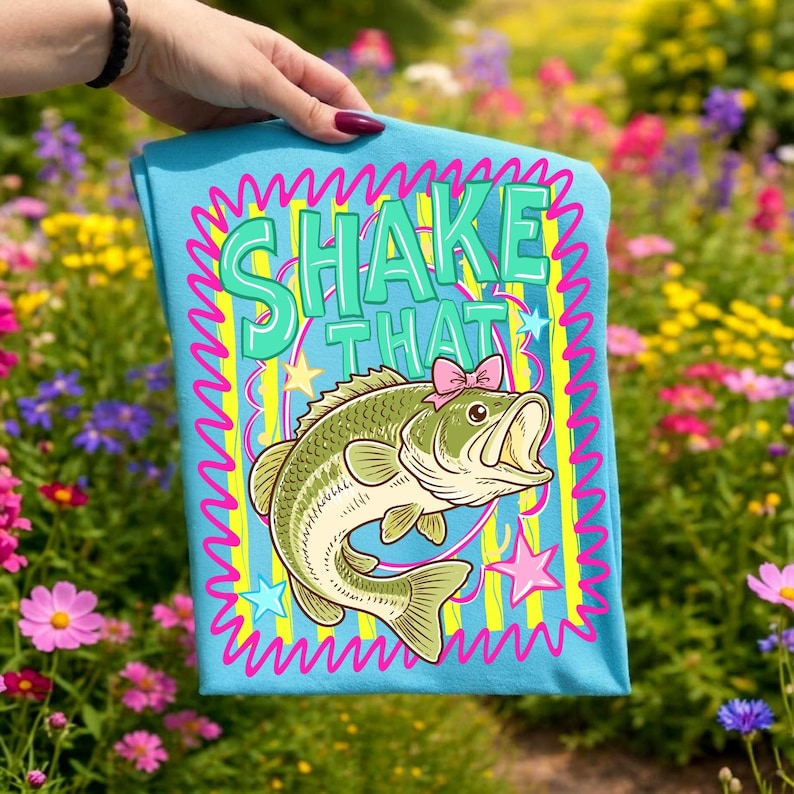 Op de afbeelding: Lichtblauw t-shirt met een cartoon bas met een roze strik en de tekst "SHAKE THAT". Het ontwerp is omlijst met een golvende roze rand en gele strepen. Het t-shirt wordt gepresenteerd voor een kleurrijke bloementuin.