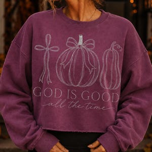 K&ouml;nnte beinhalten: Ein pflaumenfarbenes Langarm-Sweatshirt mit einer wei&szlig;en Strichgrafik, die ein Kreuz mit Schleife, einen K&uuml;rbis mit Schleife und einen Flaschenk&uuml;rbis zeigt. Der Text "GOD IS GOOD all the time" ist in Wei&szlig; unter den Illustrationen aufgedruckt.