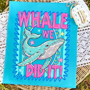 K&ouml;nnte beinhalten: Ein t&uuml;rkisfarbenes quadratisches Textil mit einer Walillustration und dem Text "WHALE WE DID IT!" in Pink. Der Wal ist hellblau mit rosa Akzenten, umgeben von Sternen und einem dekorativen Rand. Eine kleine florale Geldb&ouml;rse ist im Hintergrund.