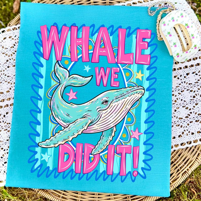 K&ouml;nnte beinhalten: Ein t&uuml;rkisfarbenes quadratisches Textil mit einer Walillustration und dem Text "WHALE WE DID IT!" in Pink. Der Wal ist hellblau mit rosa Akzenten, umgeben von Sternen und einem dekorativen Rand. Eine kleine florale Geldb&ouml;rse ist im Hintergrund.