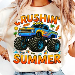 May include: Cream-colored t-shirt featuring a blue monster truck with flames and the words "Crushin' Summer" in orange and yellow. The design includes palm trees, surfboards, and beach balls.