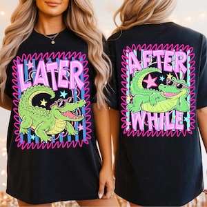 May include: Black t-shirt featuring a cartoon alligator wearing sunglasses. The front of the shirt has the word "LATER" and the alligator graphic. The back of the shirt has the words "AFTER WHILE" and the alligator graphic. The design is outlined in pink.