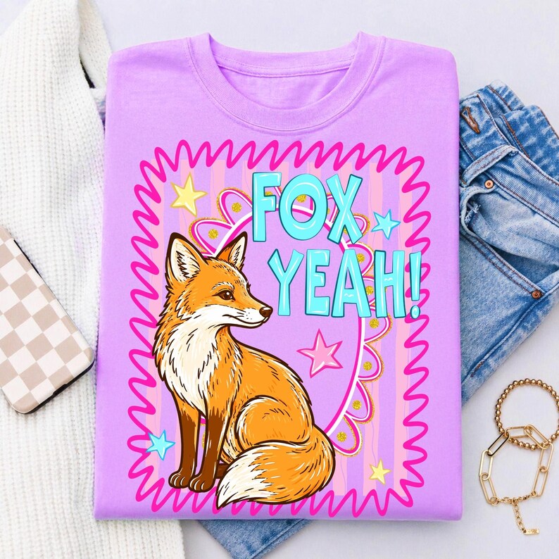 Op de afbeelding: Een lila T-shirt met een cartoon vos illustratie en de turquoise tekst "FOX YEAH!". Het ontwerp bevat een roze golvende rand, sterren en een decoratief bloemelement. De vos is oranje en wit.
