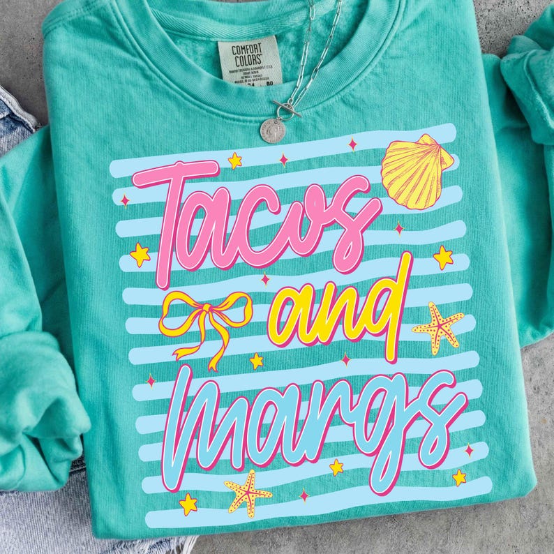 May include: Teal Comfort Colors sweatshirt with the words "Tacos and Margs" in pink and yellow, with a bow, seashells, and starfish graphics. The design is set against a background of blue and white stripes. A silver necklace is visible.