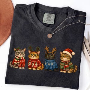 K&ouml;nnte beinhalten: Ein anthrazitfarbenes T-Shirt mit Rundhalsausschnitt und einem Aufdruck von vier Katzen in Weihnachtspullovern, Schals und Accessoires. Die Katzen tragen Brillen, Rentiergeweihe und eine Weihnachtsm&uuml;tze. Das Shirt hat ein Comfort Colors Etikett.