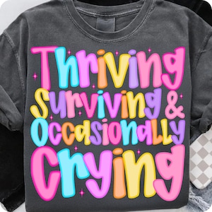 May include: Dark gray t-shirt with the words "Thriving, Surviving & Occasionally Crying" in colorful bubble letters. The text is in pink, blue, yellow, orange, and purple. The shirt is a casual style.