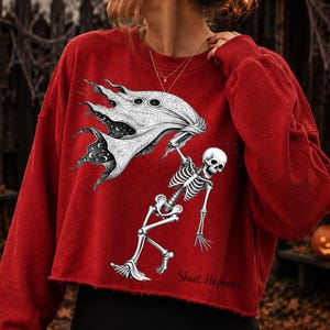 K&ouml;nnte beinhalten: Ein rotes Langarm-Sweatshirt mit offenem Saum und einem schwarz-wei&szlig;en Grafikdruck eines Skeletts, das ein Geisterlaken h&auml;lt. Das Laken zeigt ein Spinnennetz-Muster und einen Sternenhimmel. Darunter steht der Text Sheet Happens.