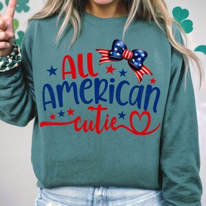 Op de afbeelding: Tealblauwe trui met een rood-wit-blauwe strik en de tekst "All American cutie" in rood en blauw.