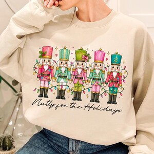 Op de afbeelding: Een beige sweater met ronde hals en een afbeelding van vijf notenkrakers die elkaars hand vasthouden, versierd met kleurrijke kerstverlichting, kleine hartjes en sterretjes. Onder de illustratie staat de tekst Nutty for the Holidays.