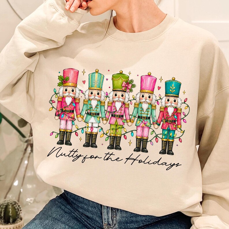 Op de afbeelding: Een beige sweater met ronde hals en een afbeelding van vijf notenkrakers die elkaars hand vasthouden, versierd met kleurrijke kerstverlichting, kleine hartjes en sterretjes. Onder de illustratie staat de tekst Nutty for the Holidays.