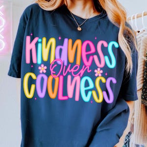 Może przedstawiać: Granatowa koszulka z napisem "Kindness Over Coolness" w kolorowych literach. Projekt zawiera odcienie r&oacute;żu, pomarańczy, ż&oacute;łci i błękitu, z małymi kwiatowymi akcentami. Codzienna, casualowa odzież.