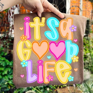 Pode incluir: Almofada decorativa castanha com a frase "it's a good life" em letras coloridas. O design inclui cora&ccedil;&otilde;es e flores, com uma fonte alegre. A almofada &eacute; segurada por uma m&atilde;o, criando um ambiente positivo.