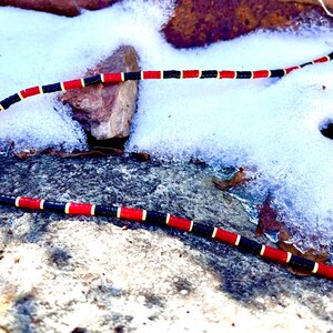 May include: A beaded necklace with a pattern of red, black, and yellow beads. The necklace is lying on a stone surface covered in snow. The necklace has a silver-coloured clasp.