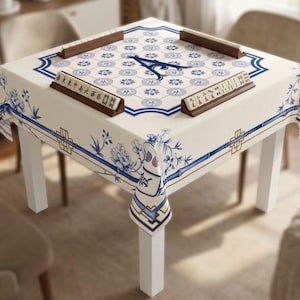 May include: A square table covered with a cream-colored tablecloth featuring blue floral patterns and geometric borders. The tabletop displays a mahjong set with tiles arranged in wooden racks and a central letter A on a grid of floral motifs.