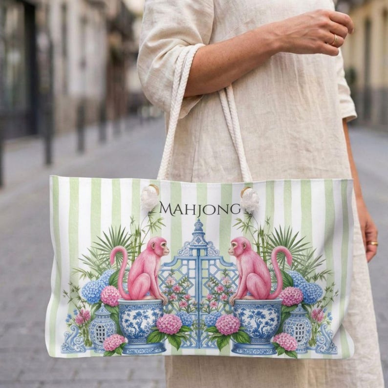 May include: A tote bag featuring a light green and white vertical striped pattern. The design includes two pink monkeys sitting on blue and white patterned ceramic pots, flanking a central blue gate, surrounded by green foliage and pink flowers.
