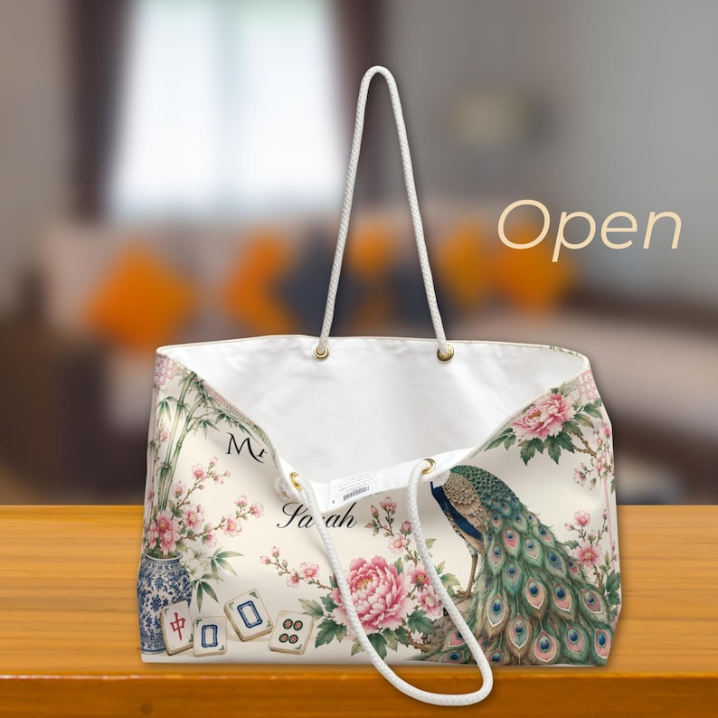 May include: A cream-colored tote bag with white rope handles featuring a printed design of a peacock, pink peonies, bamboo, and mahjong tiles. The text "Sarah" is printed on the front, and the word "Open" appears in the upper right corner.