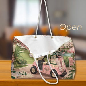 May include: A large tote bag with white rope handles featuring a printed scene of a pink golf cart on a course, a mahjong set on a table, and lush greenery. The interior is white, and the word Open is printed in the upper right corner of the image.
