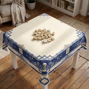 May include: A square tablecloth with a cream-colored center and a wide blue border featuring bamboo, floral, and geometric patterns. A collection of mahjong tiles is arranged in the center of the table, displaying various Chinese characters and symbols.