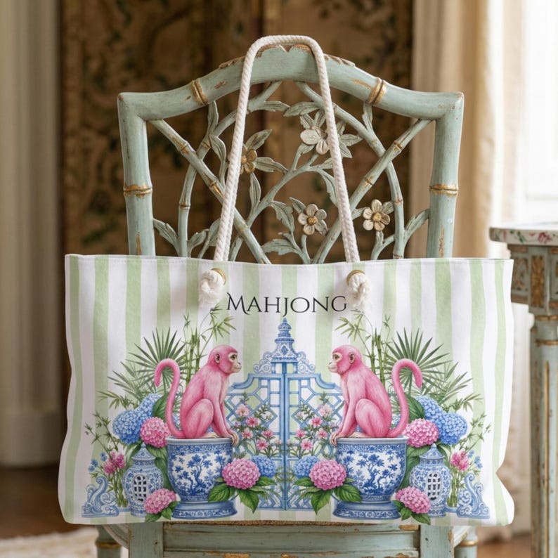 May include: A white and light green striped tote bag with rope handles. The front features a graphic of two pink monkeys sitting on blue and white patterned urns, flanking a blue gate, with the word MAHJONG printed in black above the gate.