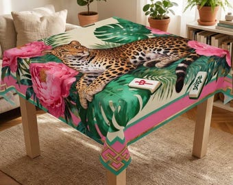 Puede incluir: Un mantel cuadrado con un diseño de estampado de leopardo, que presenta un leopardo descansando sobre un patrón de hojas y flores tropicales. El mantel tiene bordes rosas y verdes e incluye dos fichas de Mahjong. La mesa es de madera.