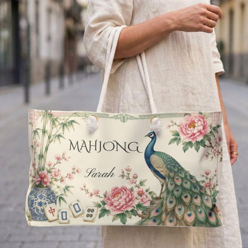 May include: A cream-colored tote bag featuring a peacock, pink peony flowers, bamboo stalks in a blue and white vase, and mahjong tiles. The text "MAHJONG" and the name "Sarah" are printed in black script and serif fonts on the front of the bag.