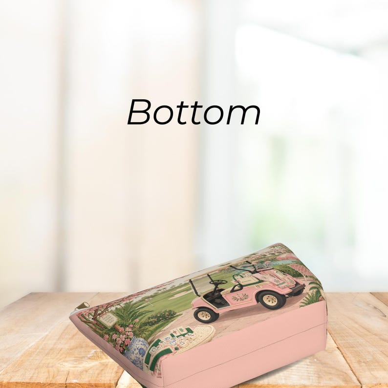May include: A pink cosmetic pouch featuring a printed graphic of a pink golf cart on a green course with mahjong tiles in the foreground. The word Bottom is printed in black text above the pouch. The pouch has a silver zipper closure.