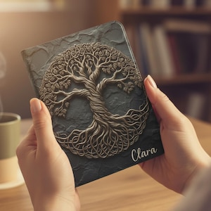 May include: A dark gray journal with a detailed, raised tree of life design in a metallic bronze color. The name "Clara" is printed in white script on the lower right corner. The journal is held in two hands.