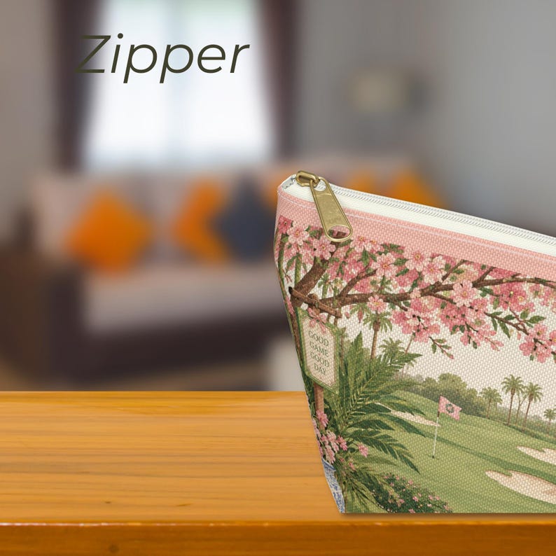 May include: A zippered pouch featuring a golf course landscape with green fairways, sand traps, palm trees, and pink cherry blossom branches. The fabric has a light beige background and a gold-toned metal zipper pull. The text GOOD GAME GOOD DAY is visible.