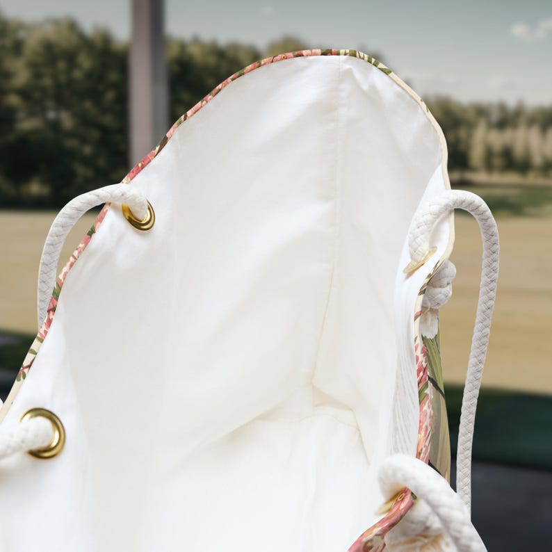 May include: A tote bag with a white interior lining and a floral exterior pattern featuring pink and green tones. The bag includes thick white rope handles attached through gold-toned metal grommets, providing a sturdy structure for the open-top design.