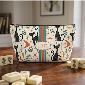 May include: A zippered pouch with an off-white background featuring a repeating pattern of black cats, orange boomerangs, green leaves, and starbursts. A central vertical stripe contains a label with the word TILES in red lettering.