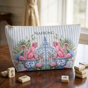 May include: A zippered pouch with a light blue and white vertical striped pattern. The front features two pink monkeys sitting in blue and white patterned pots, flanking a central gate, with floral accents and the word MAHJONG printed above.