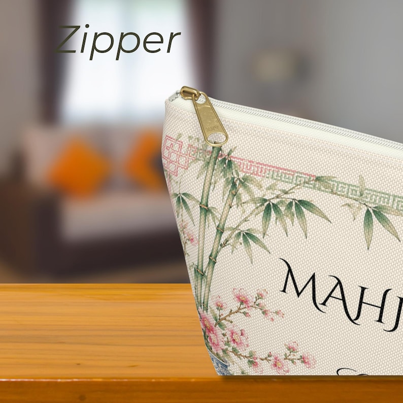 May include: A beige fabric pouch featuring a gold-tone metal zipper and a printed design of green bamboo stalks, pink cherry blossoms, and a geometric border. The text "MAHJ" is visible on the front, along with the word "Zipper" at the top left.