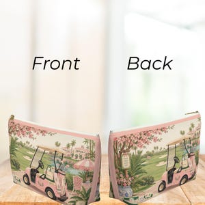 May include: A rectangular zippered pouch featuring a pink golf cart on a golf course with palm trees and a building in the background. The design includes a pink and white striped umbrella, a floral tree, and a sign that reads Good Game Good Day.