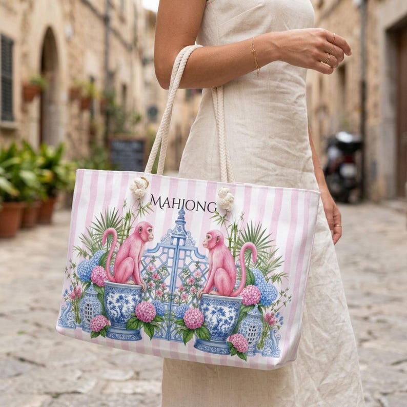 May include: A tote bag with a light pink and white vertical striped pattern. The front features the text MAHJONG above two pink monkeys sitting on blue and white patterned ceramic vessels, surrounded by blue gates and pink and blue hydrangea flowers.